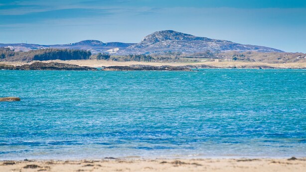 Paesaggio costiero con acque turchesi cristalline, spiaggia sabbiosa e colline rocciose in lontananza.