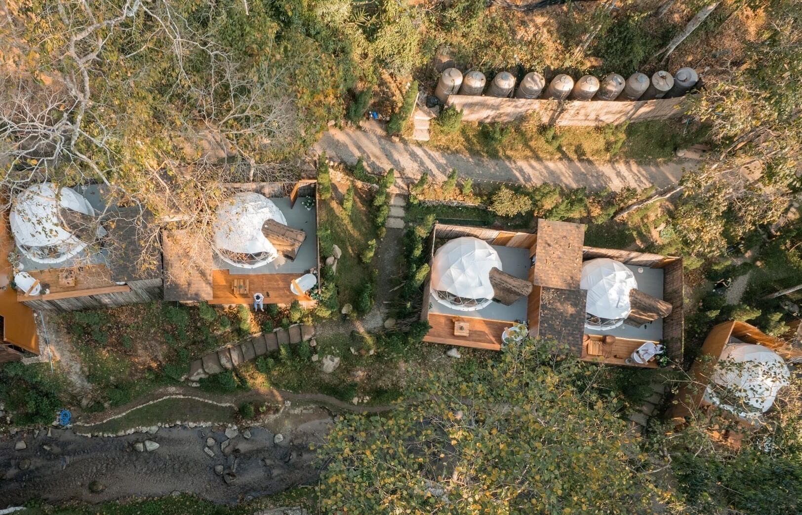 Vista aérea de una cabaña rústica con techo de paja, rodeada de un bosque frondoso. Se observan tres cabañas con terrazas de madera y estructuras circulares blancas, posiblemente para comer o relajarse.