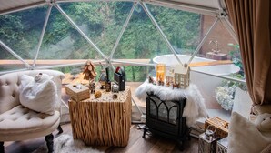 Una habitación acogedora con un ventanal grande, una mesa de madera, un sillón cómodo, una chimenea y vistas al bosque.