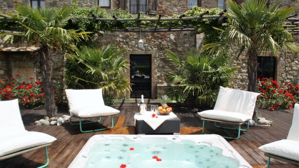 Un jacuzzi con sillas blancas y una mesa, rodeado de follaje y un muro de piedra.
