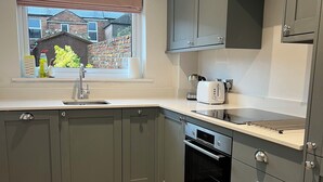 A modern kitchen with grey cabinets, a built-in oven, and a window with a view of a brick house. - Train Station Cottage Taunton - Walk to town, pet friendly! (Taunton)