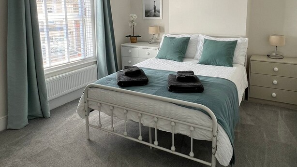 A bedroom with a white bed, teal bedding, a white radiator, a window with curtains, a bedside table with a lamp, and a picture on the wall. - Train Station Cottage Taunton - Walk to town, pet friendly! (Taunton)