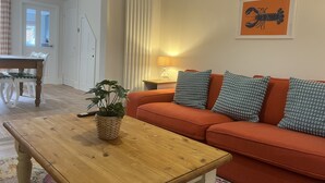 A living room with a wooden coffee table, an orange sofa with patterned cushions, a potted plant, and a framed artwork on the wall. - Train Station Cottage Taunton - Walk to town, pet friendly! (Taunton)