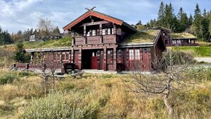 A traditional wooden house with a balcony, set in a grassy area with trees in the background.