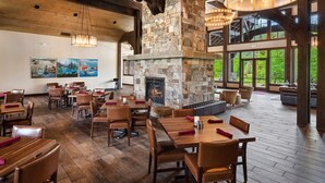 A spacious dining area with wooden tables and chairs, a stone fireplace, and large windows offering views of the outdoors.