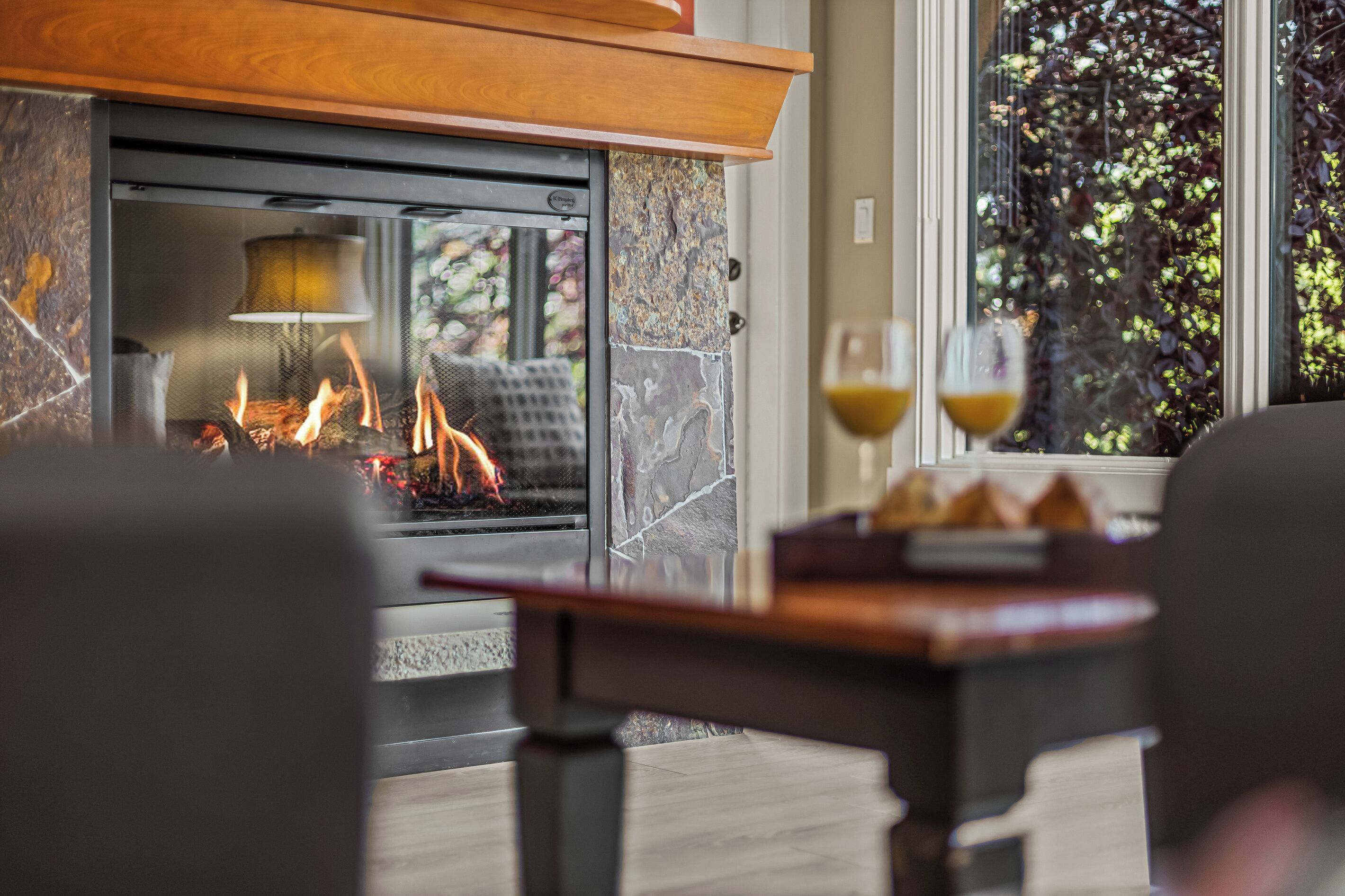 A cozy indoor setting with a fireplace, a table with glasses, and a view of greenery outside.