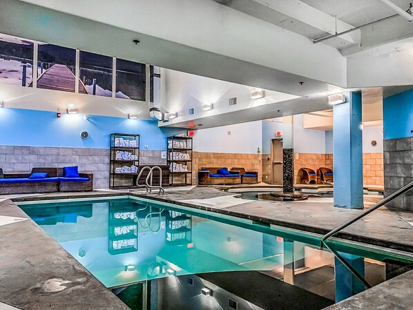An indoor swimming pool with lounge areas, towels on shelves, and a view of the outside.