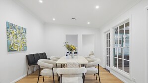 Comedor con una mesa blanca, seis sillas, un sofá negro y un ventanal con contraventanas blancas.