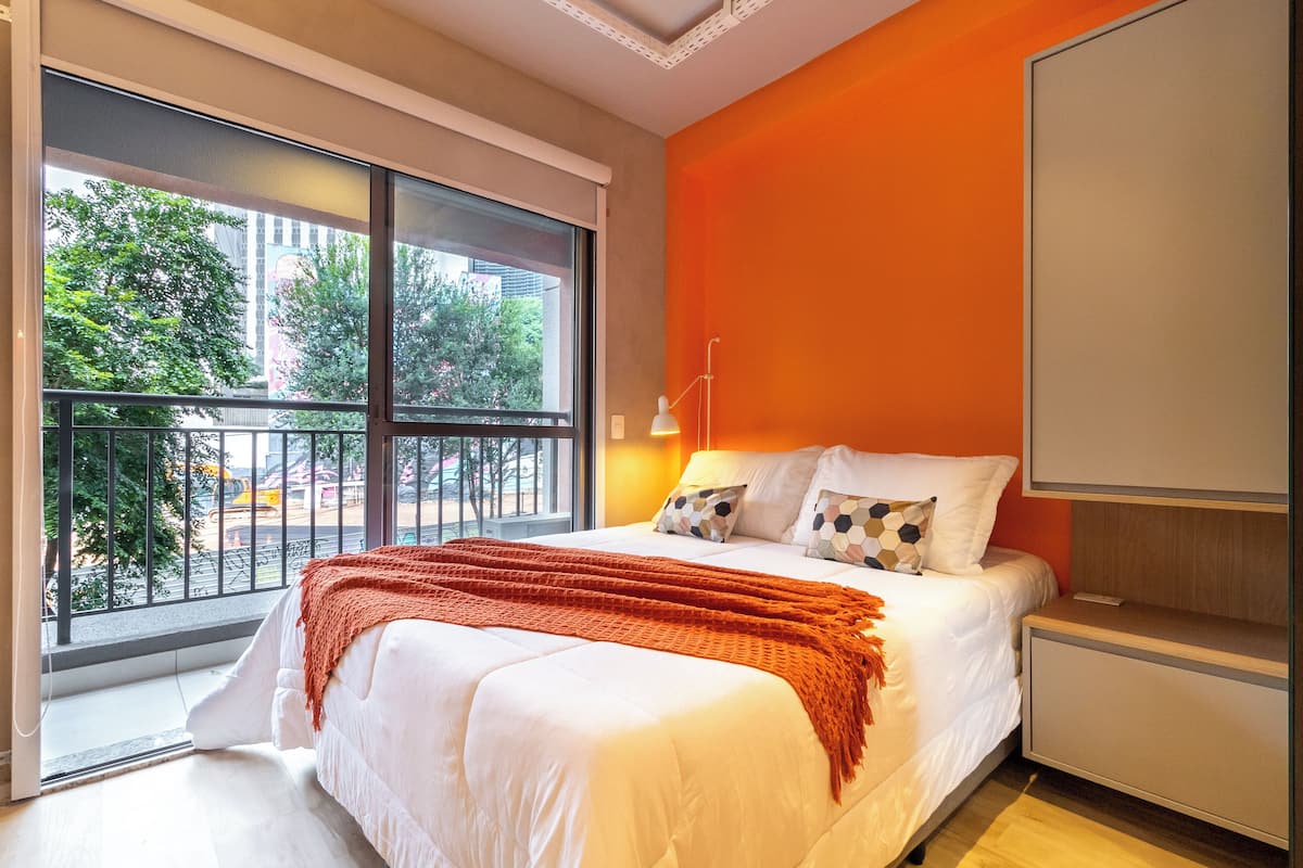 A modern hotel room with a large window, a bed with white bedding and orange accents, and a bright orange accent wall.