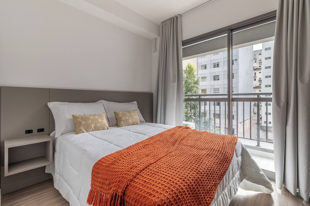 A modern bedroom with a large bed, a nightstand, and a view of a cityscape through a balcony.