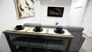 Et spiseområde med et træbord og fire sorte skåle på underlayere, en grå sofa og et Disney+-skilt monteret på væggen.