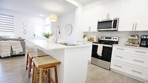 A modern kitchen with white cabinetry, a stainless steel oven, and a built-in microwave. There are wooden stools at the island, and a sink with a faucet. - BoHo Retreat with King Beds|A/C|Disney+|Near WEM (Edmonton)