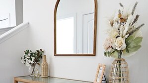 Una consola de madera con un espejo encima, una planta pequeña y un jarrón decorativo con flores.