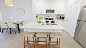 Una cocina moderna con armarios blancos, una isla central con taburetes, un comedor con sillas y una lámpara colgante.