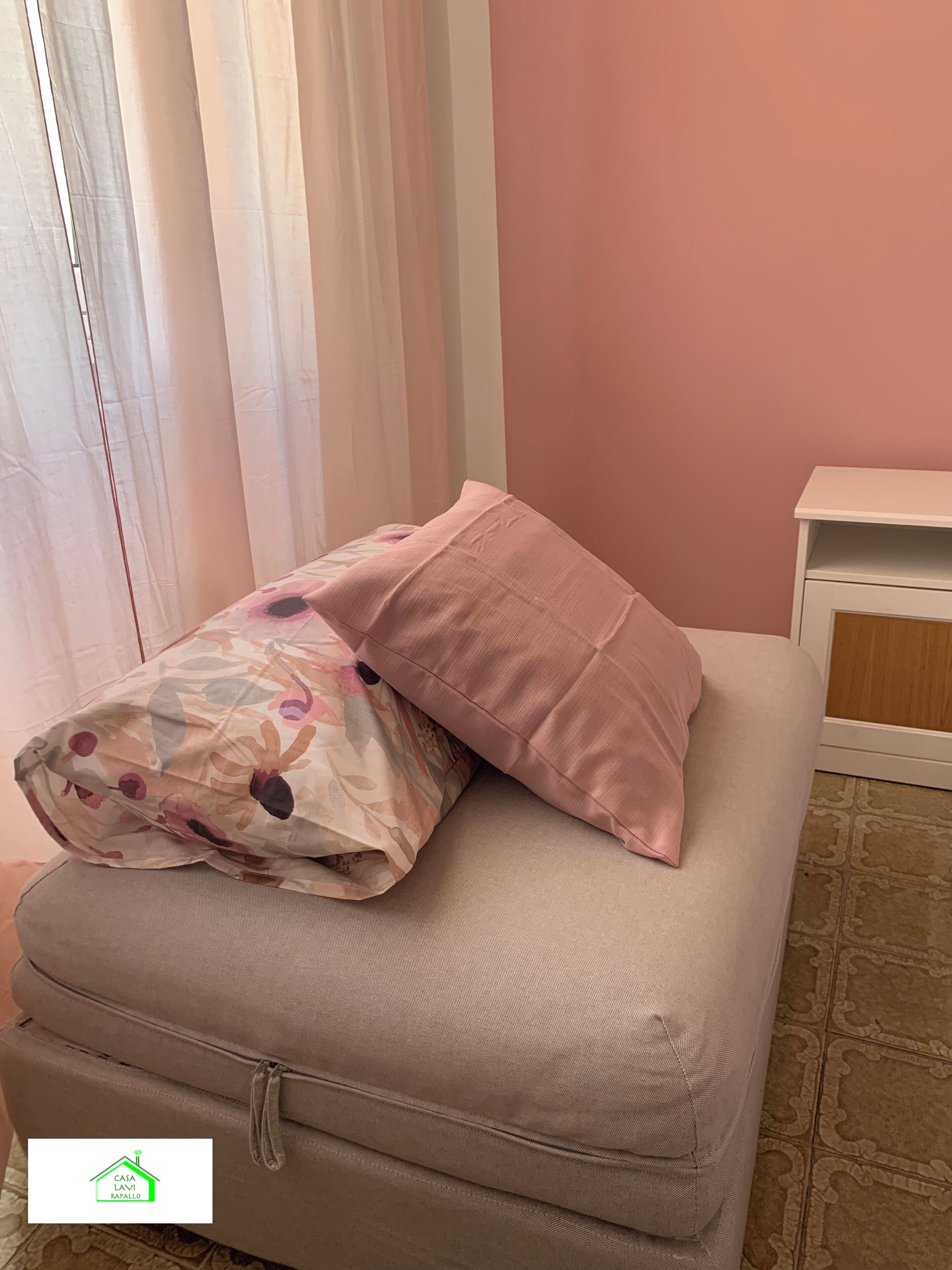 Ein Bett mit einer decke mit floralem Muster und einem rosafarbenen Kissen, ein kleiner Nachttisch und eine Wand in Rosa.