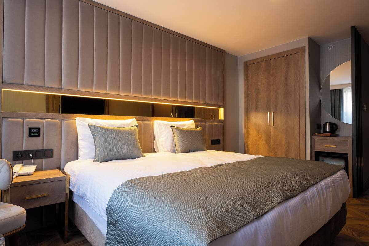 A modern hotel room with a large bed, a bedside table with a lamp, a built-in TV, and a wardrobe.