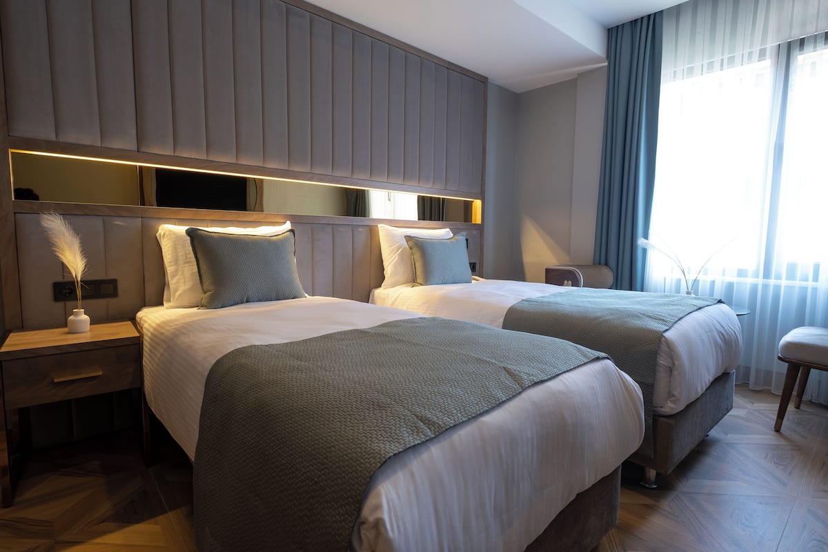 A hotel room with two beds, a nightstand, a bedside table, a mirror, and a window with curtains.