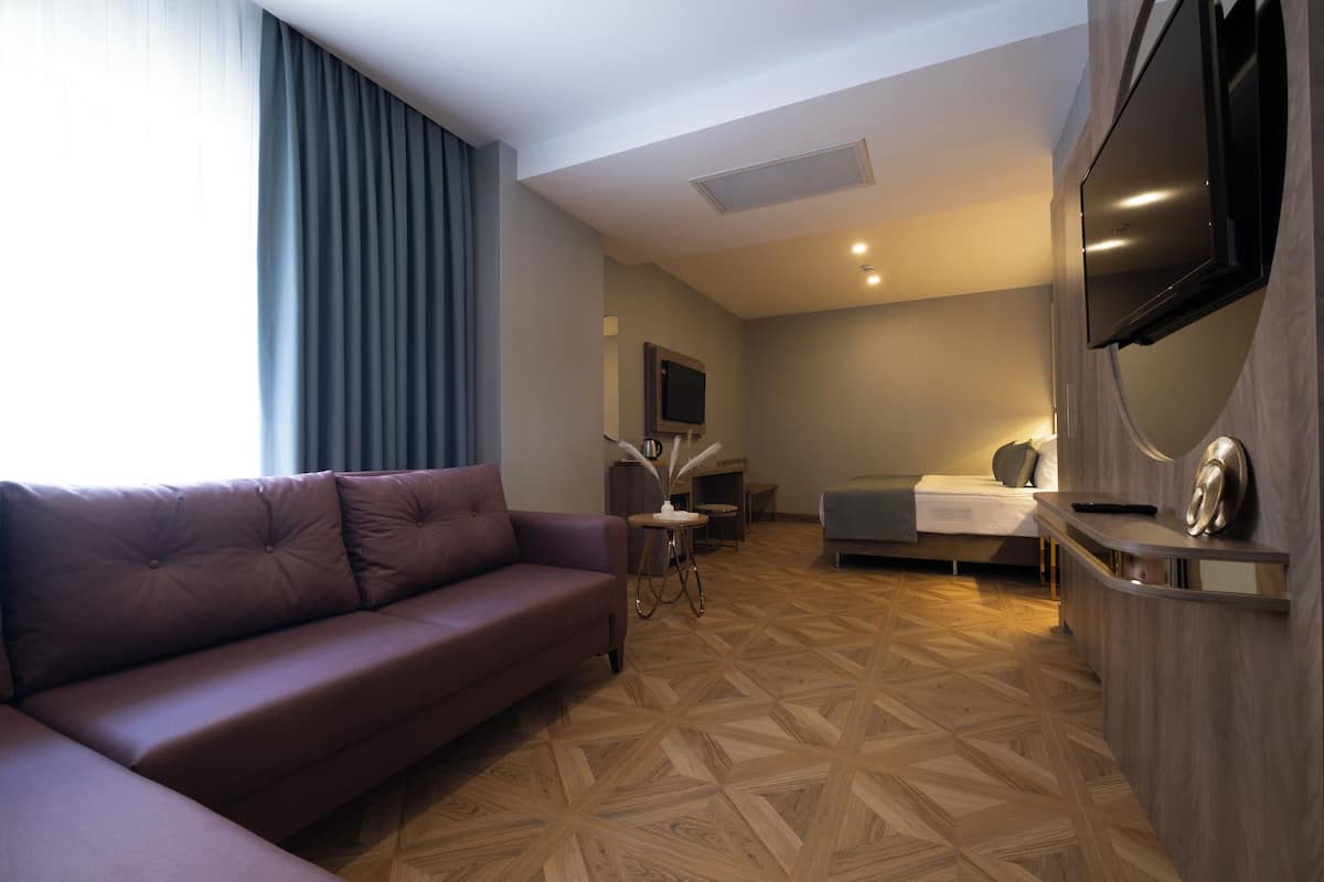 A modern hotel room with a brown leather sofa, a bed with a gray headboard, a flat-screen TV mounted on the wall, and a wooden parquet floor.