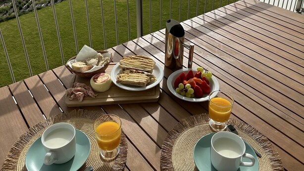 Desayuno dispuesto en un balcón con vistas a un paisaje pintoresco que incluye un puente y una playa.