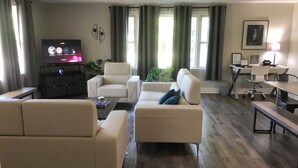 Een moderne woonkamer met een flatscreen televisie, comfortabele zithoeken, een bureau en een raam met gordijnen.