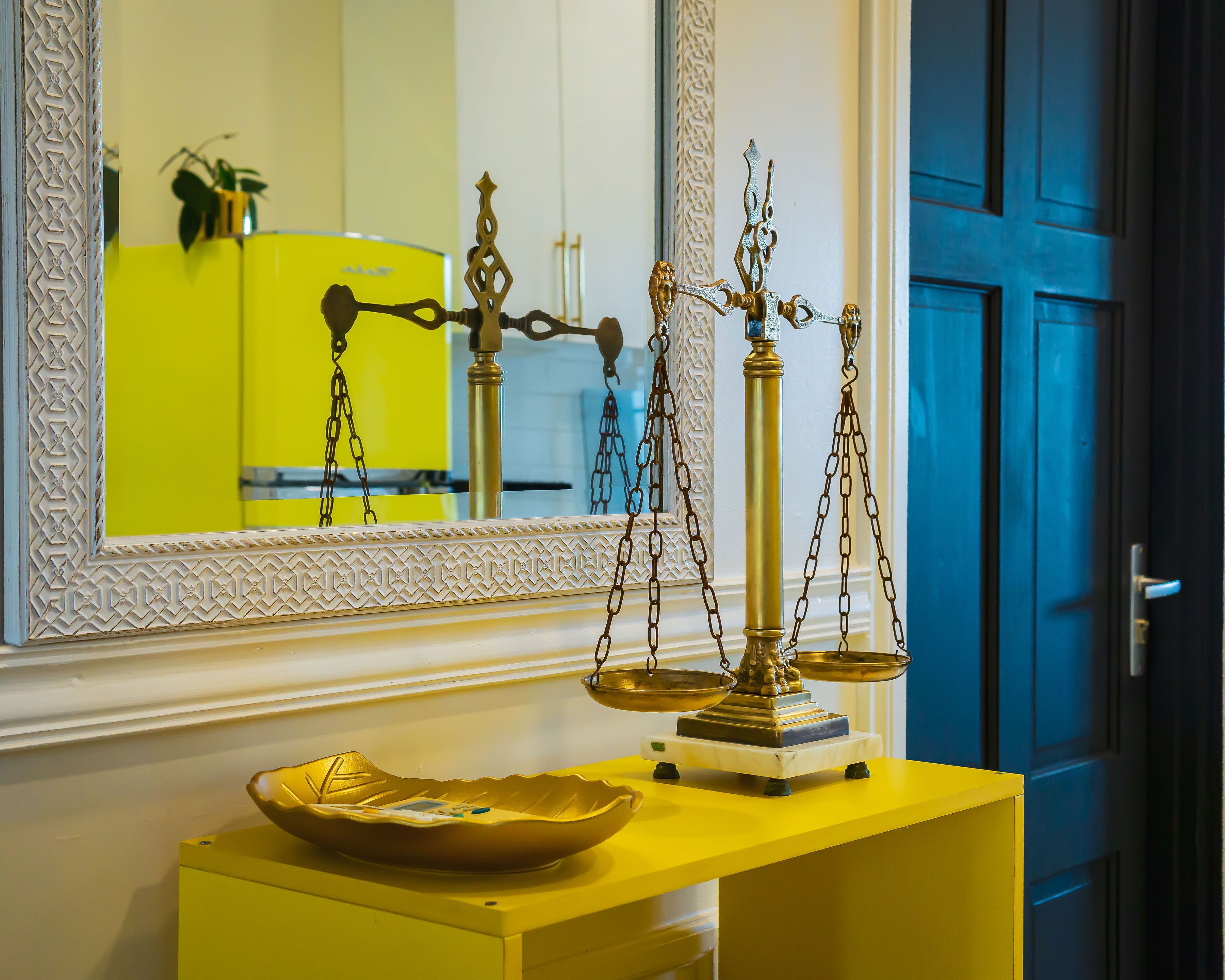 A scale on a yellow console table with a decorative bowl on it, a mirror with a geometric border, and a blue door in the background.