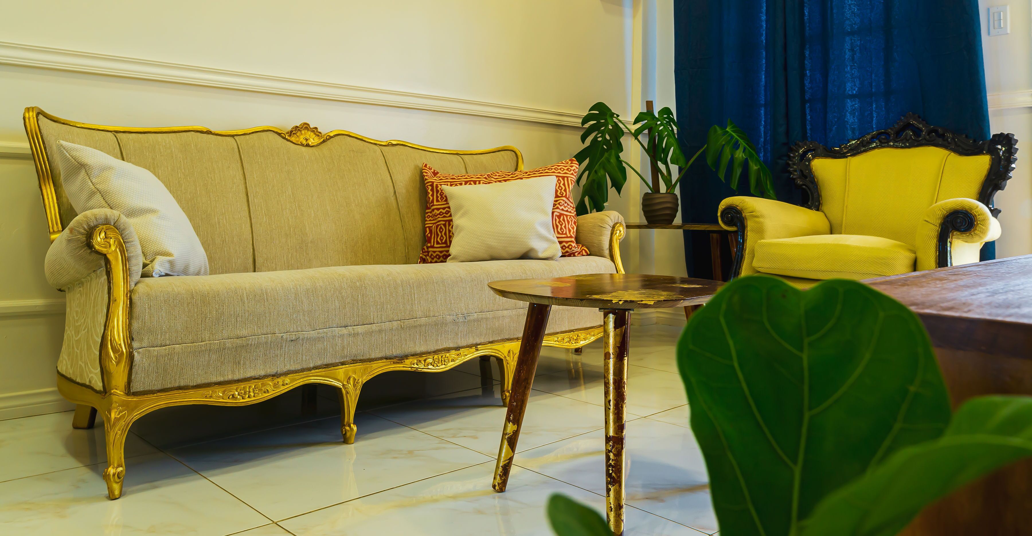 A room with a beige sofa, a yellow armchair, and a small table. There are two potted plants and a blue curtain in the background.