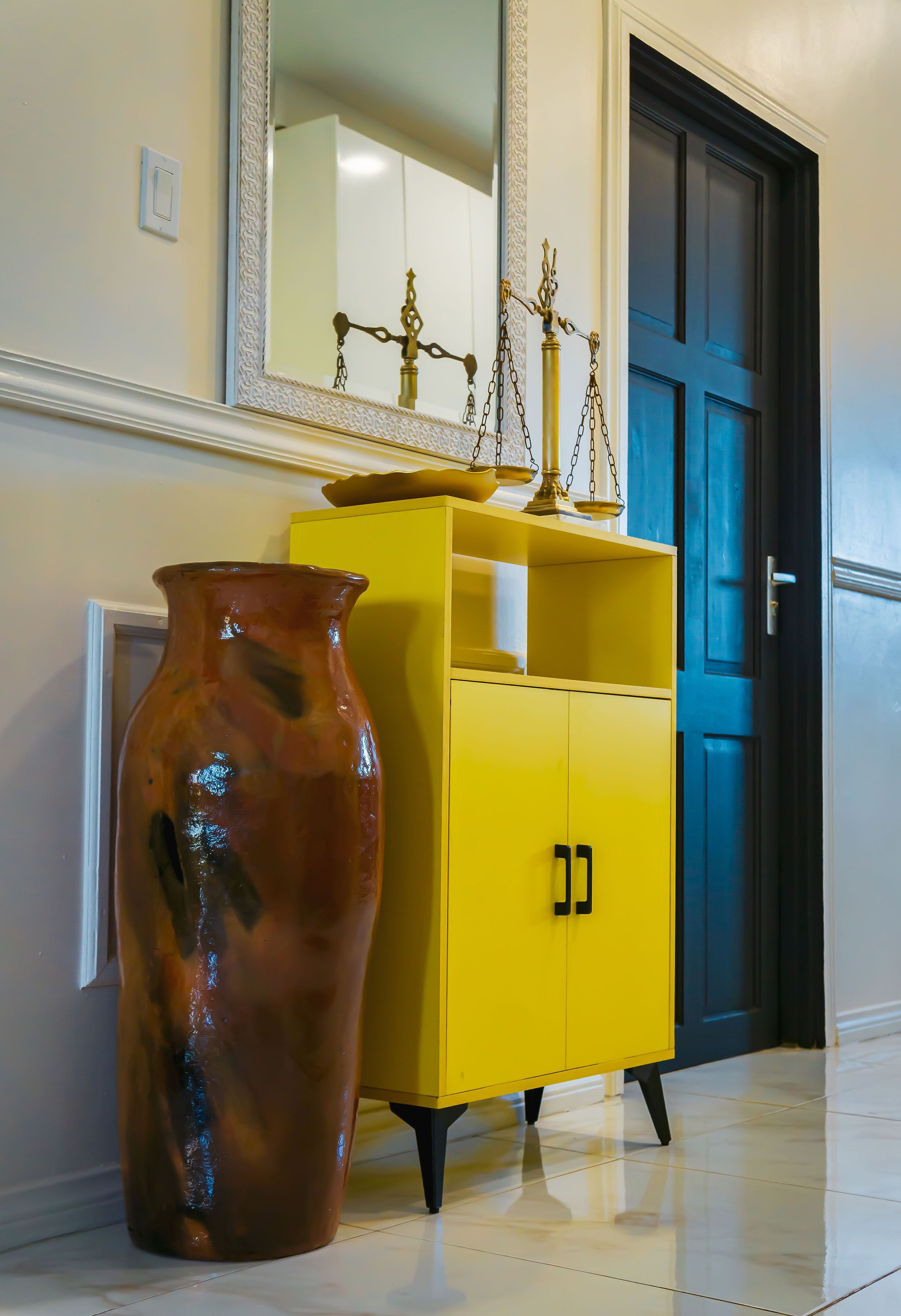 Un mueble amarillo con herrajes negros, un jarrón grande y un espejo encima.