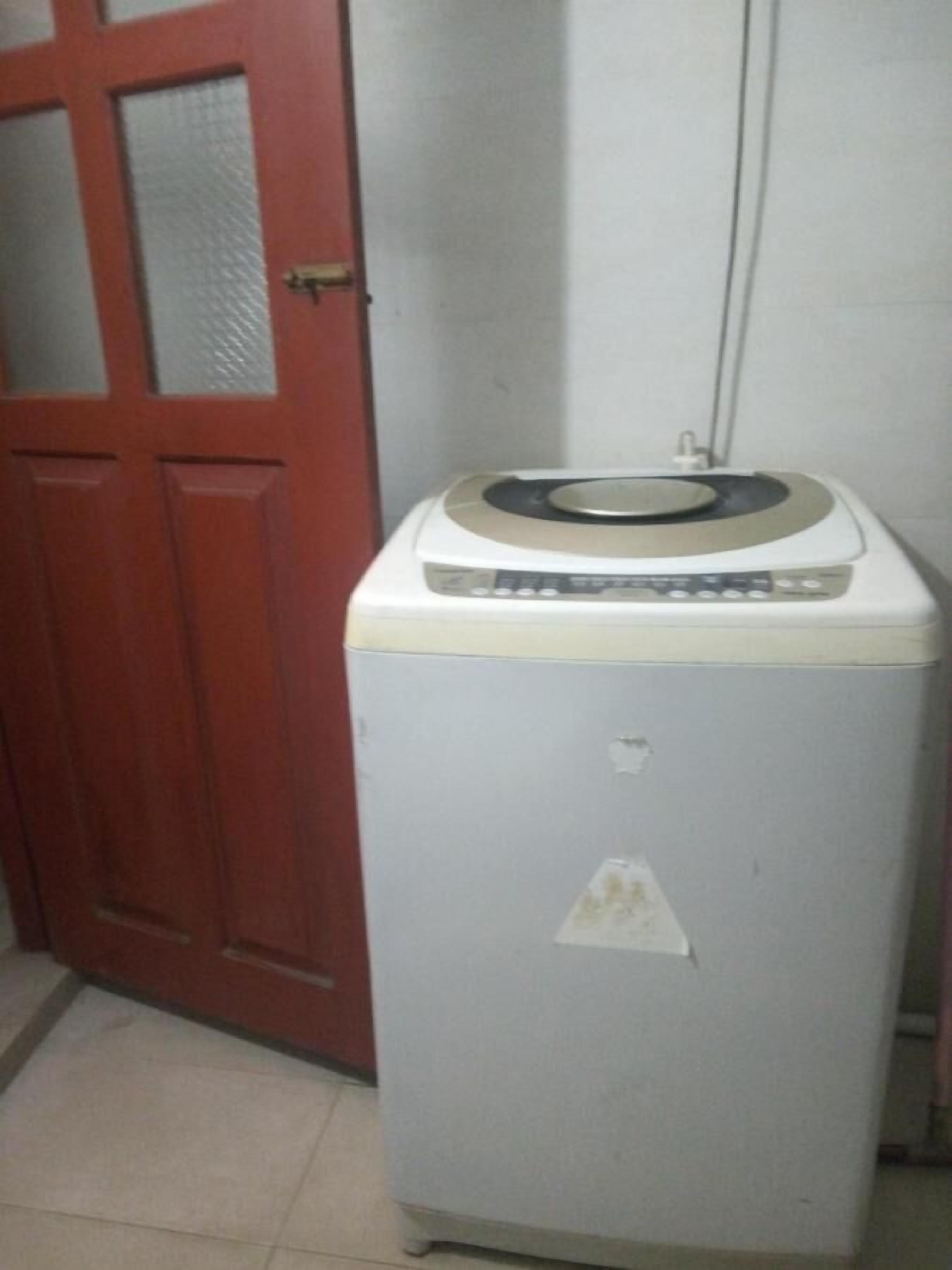 A compact, white washing machine with a control panel, placed in a room with a red door and tiled floor.