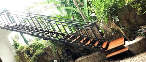 A staircase with metal railings and wooden steps, surrounded by greenery and a stone wall.
