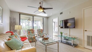 Ein Wohnzimmer mit Glastisch, einer Couch mit gemusterten Kissen, Deckenventilator und einem an der Wand befestigten Fernseher.
