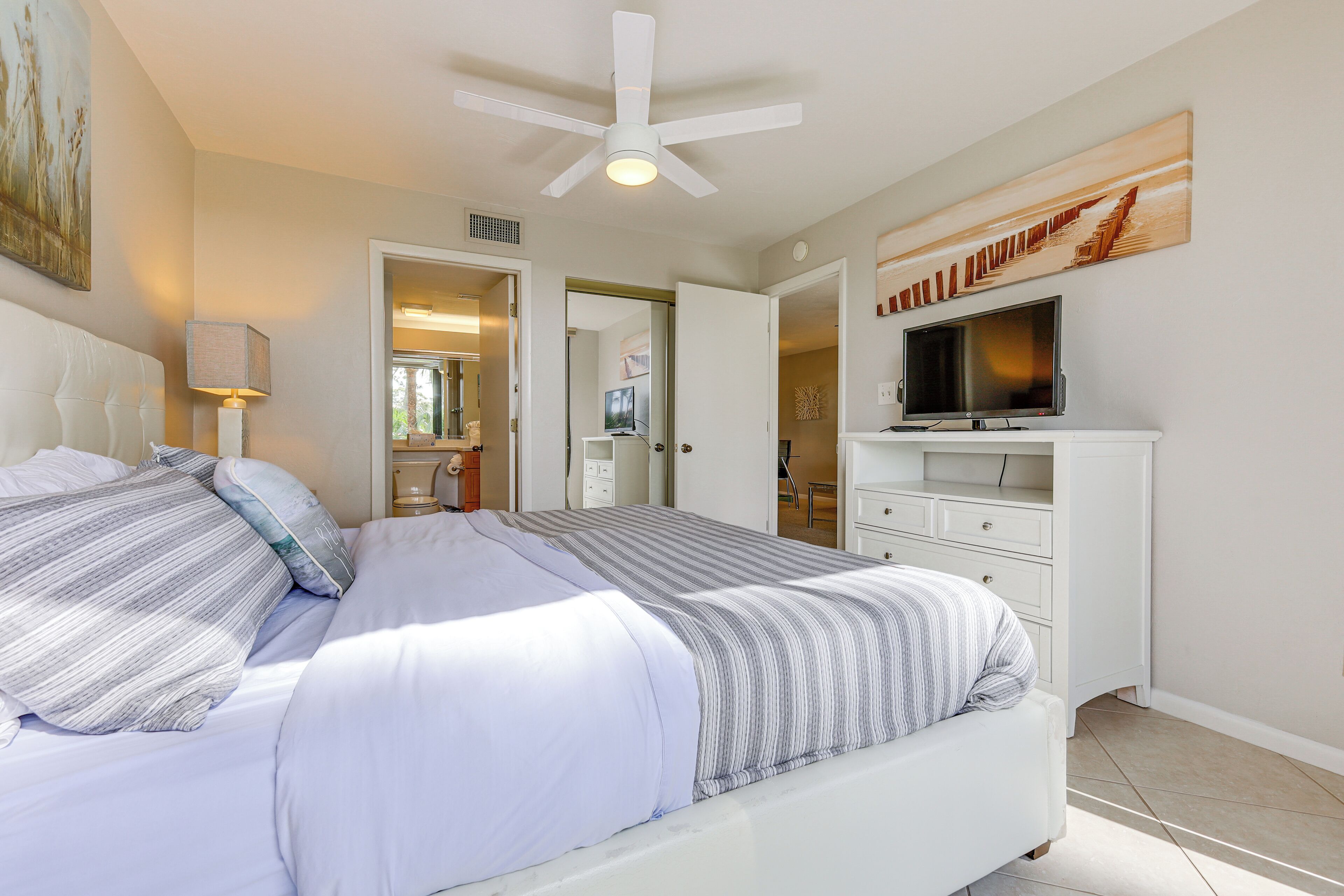 A bedroom with a bed, a ceiling fan, a TV, and a painting on the wall.
