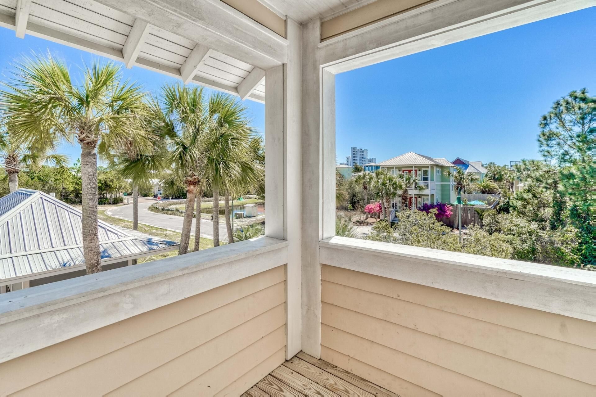 A covered balcony with a view of a residential area, palm trees, and a house with a red roof.