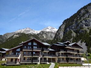 A multi-story hotel with balconies, set against a backdrop of snow-capped mountains and lush greenery.