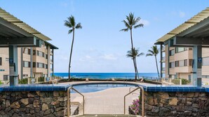A pool area with a stone wall, palm trees, and a view of the ocean. - Big Island Kona Reef D-22 3 Bedroom Condo (Kailua-Kona)