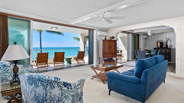 A spacious living area with a large window overlooking a balcony with a view of the ocean. There is a blue sofa, a wooden coffee table, and a lamp. - Big Island Kona Reef D-22 3 Bedroom Condo (Kailua-Kona)