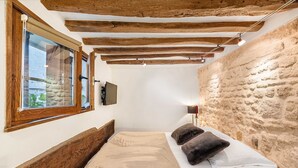 A bedroom with a wooden headboard, a large window with blinds, a stone wall, and a bedside lamp. - Aquipic, 2BR/1BA, 4 people (París)