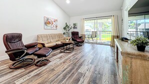 Uma sala de estar espaçosa com um sofá e uma poltrona de couro marrom, e uma mesa de centro de madeira. Há uma janela grande com vista para uma paisagem verde.