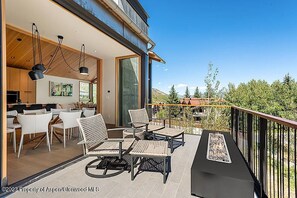 A modern outdoor patio with wicker furniture, a dining area with white chairs, and a kitchen visible through large windows.