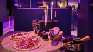 Un comedor con una mesa dispuesta para dos personas, que incluye una selección de quesos, carnes y frutas. Hay velas y una botella de champán sobre la mesa. La habitación presenta un diseño moderno con iluminación púrpura.