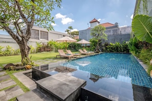 A swimming pool with lounge chairs and umbrellas, surrounded by greenery and a building in the background. - Tanaka Canggu Guesthouse (Canggu)