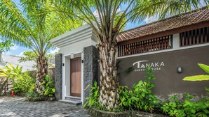 Rakennuksessa on kyltti, jossa lukee ”Tanaka Guest House”, sen edessä on asfaltoitu sisäänkäynti, ja sitä ympäröi vehreää kasvillisuutta.
