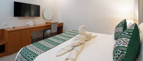 A hotel room with a bed, a desk, a chair, a flat-screen TV, a round mirror, and patterned pillows.