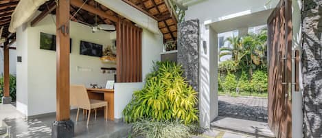 A covered outdoor area with a wooden table and chairs, a stone wall, and a large plant.