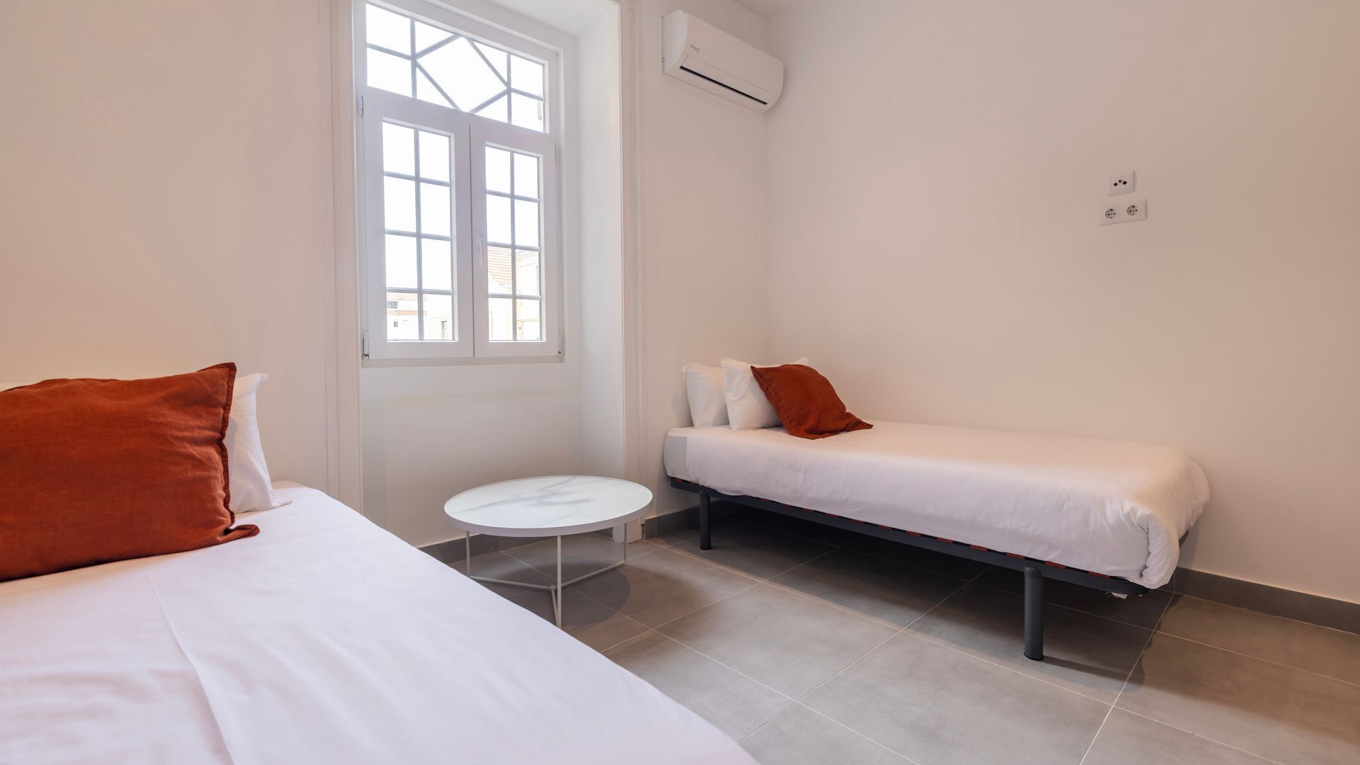 A room with two beds, a small round table, a window with white shutters, and an air conditioning unit on the wall.