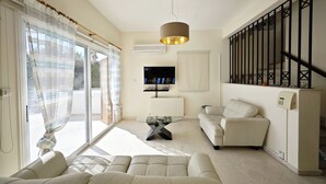 Ein modernes Wohnzimmer mit einer Couch, einem Couchtisch und einem Fernseher. Rechts befindet sich eine Treppe mit Geländer, links ein Fenster mit Vorhängen.