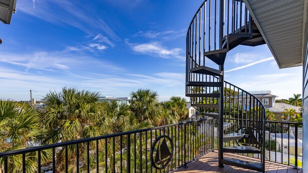 A spiral staircase on a wooden deck with a view of palm trees and a building in the background.