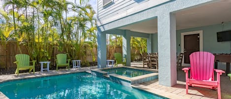 A swimming pool with a wooden deck, green and pink Adirondack chairs, and a covered patio area.