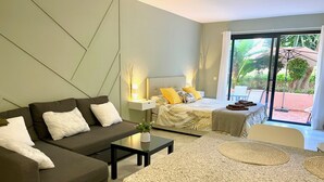 A modern hotel room with a sofa, a bed with a tropical-themed bedspread, a small table with a vase, and a large window with a view of greenery. - Banus Elegant Modern Estudio Next to the Beach (Marbella)