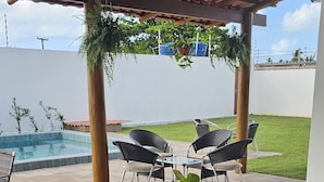 Un espace de restauration en plein air avec vue sur la piscine, comprenant une table en bois, des chaises noires et un verre de jus d’orange.