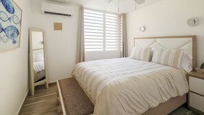 Ein Schlafzimmer mit Bett, Deckenventilator, Fenster mit Jalousien, Spiegel und Wandklimaanlage.